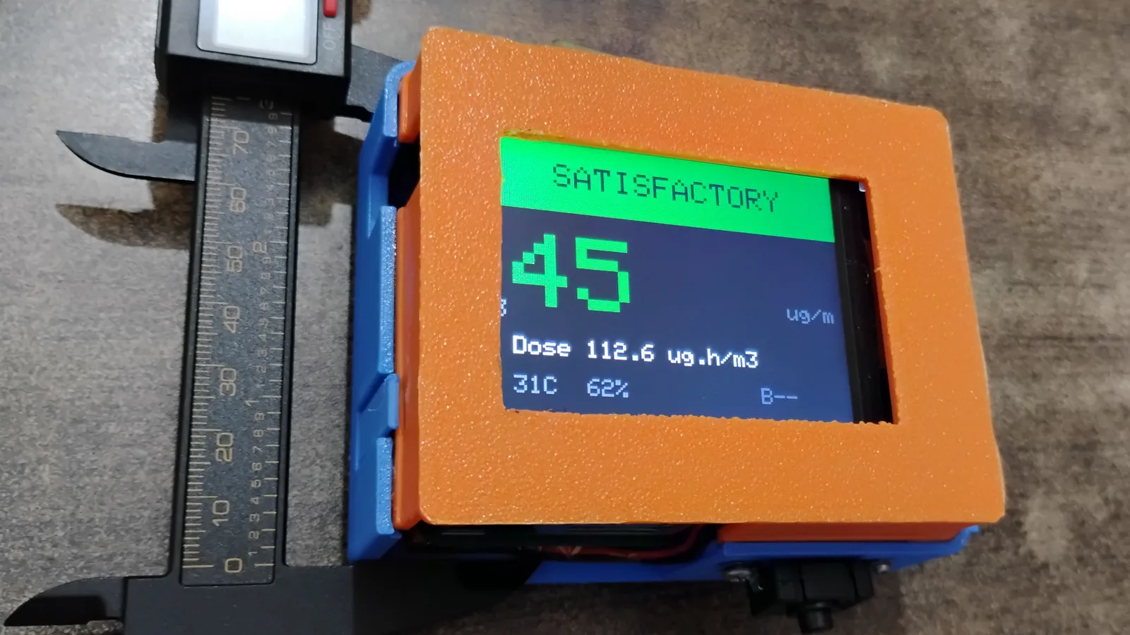 Aether badge prototype in an orange and blue 3D-printed enclosure, its display showing Satisfactory, 45 µg/m³ PM2.5, a shift dose of 112.6 µg·h/m³, 31 °C and 62% humidity, beside a caliper for scale